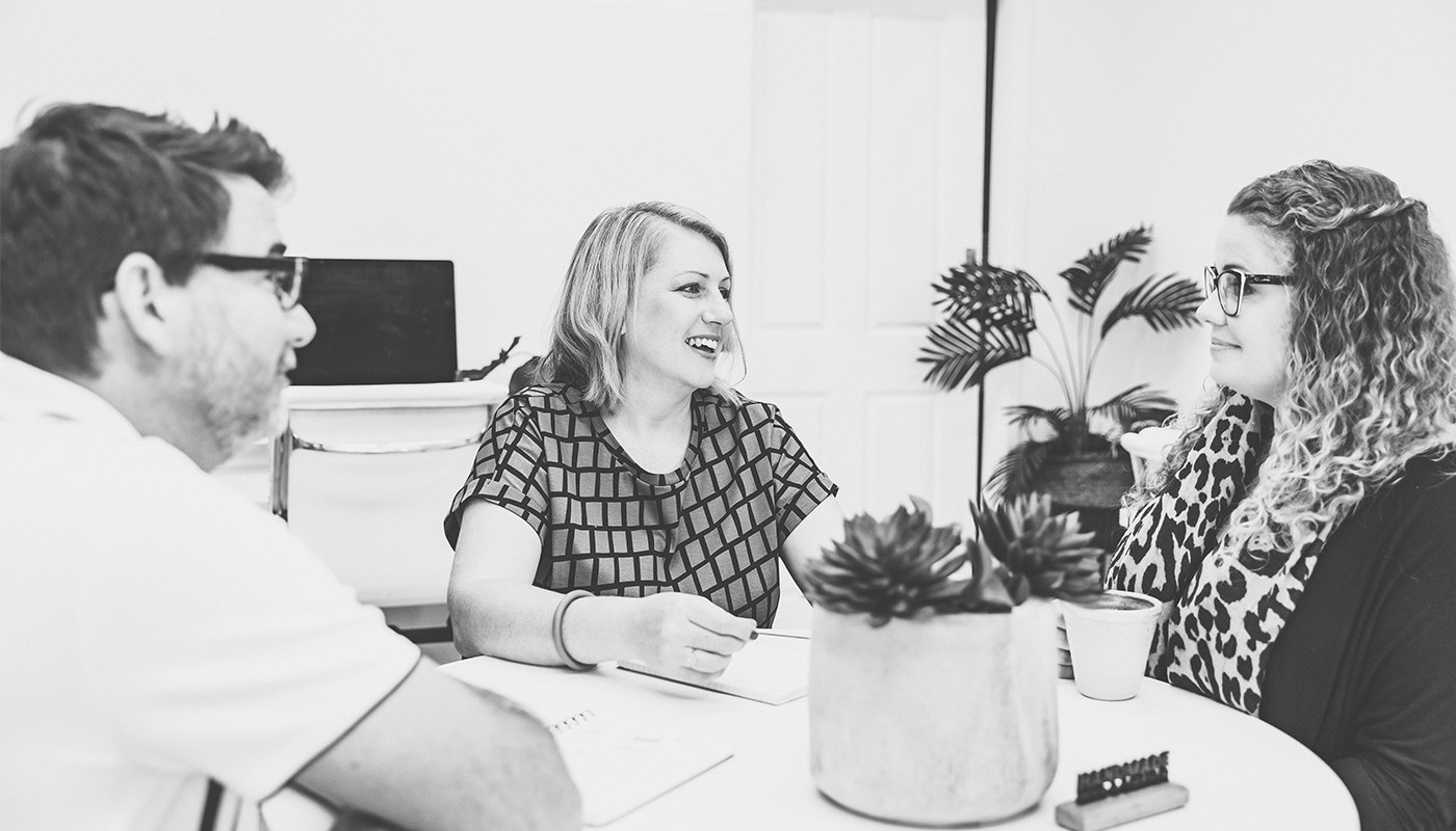 A black and white image of three people sitting around a table talking with notepads, engaged in conversation